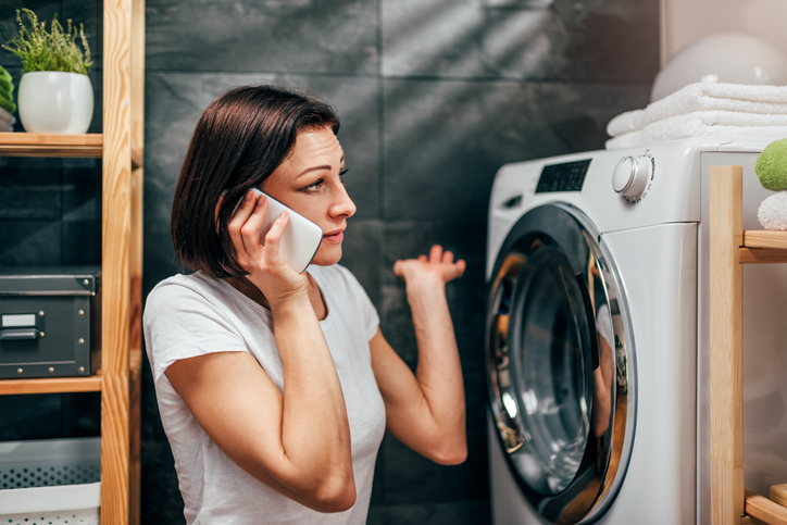 Woman wearing white shirt sitting at landry room by the washing machine and calling for appliance repair service